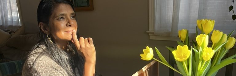 A person with long hair and a thoughtful expression sits at a table, resting their chin on their hand. In the foreground, a vase of yellow tulips is visible, illuminated by natural light from a nearby window.